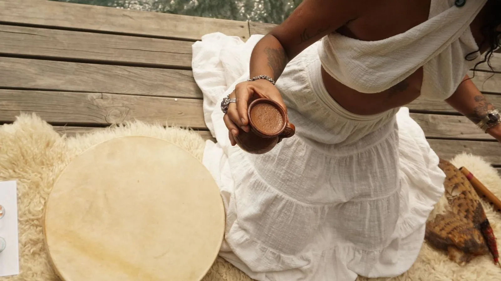 Pessoa vestindo branco segurando uma caneca marrom com bebida cremosa, sentada em tapete peludo sobre piso de madeira.