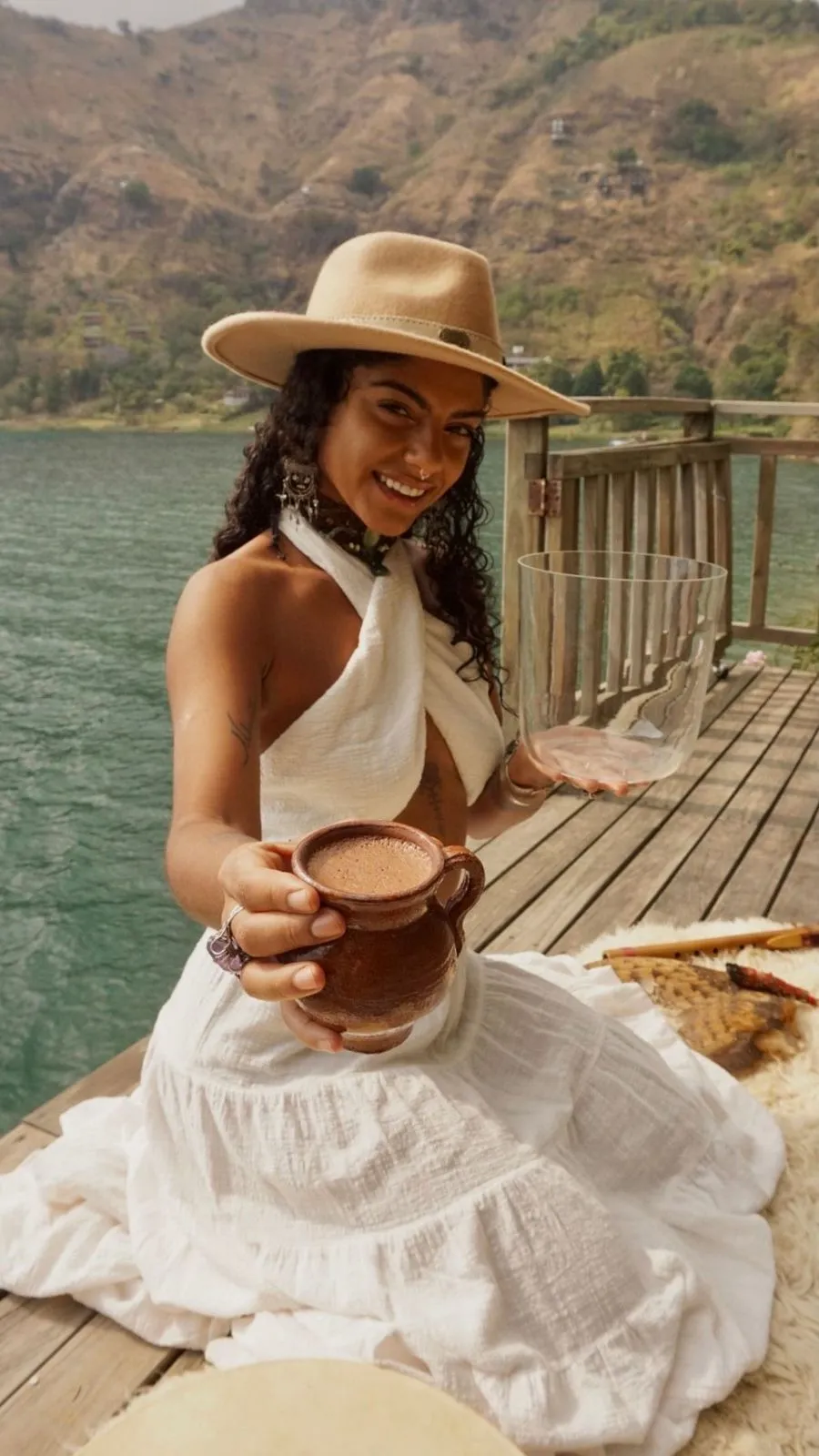 Mulher sorridente com chapéu bege e vestido branco oferece uma caneca de barro com bebida, sentada em um deque de madeira próximo a um lago com montanhas ao fundo.