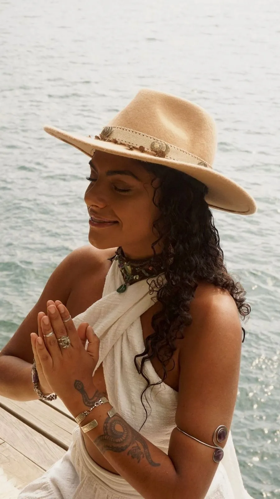 Mulher de chapéu bege meditando com as mãos em posição de oração perto da água.