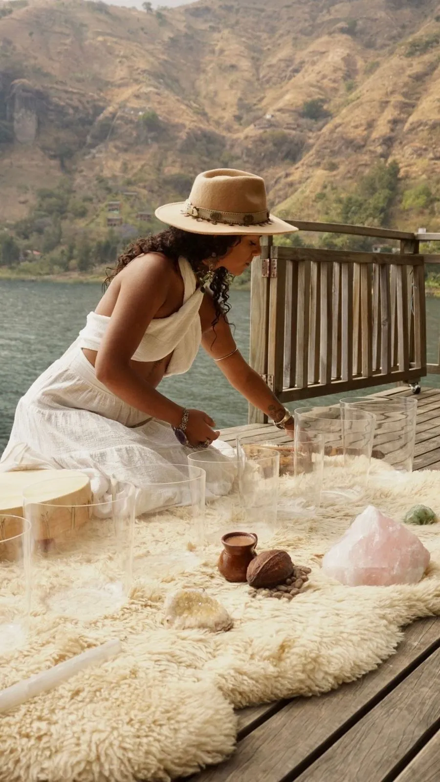 Mulher de vestido branco e chapéu bege sentada em deck de madeira ao lado de cristais e instrumentos musicais, com montanhas e lago ao fundo.