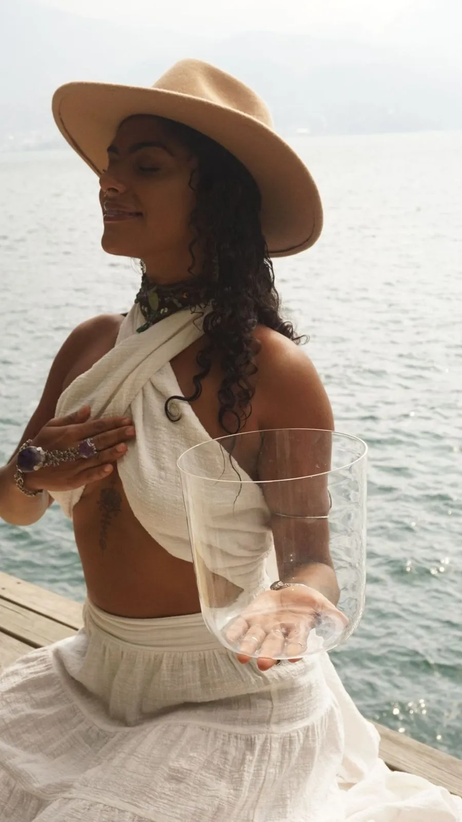 Mulher de chapéu bege e roupa branca segurando um copo de vidro transparente à beira de um corpo d'água.