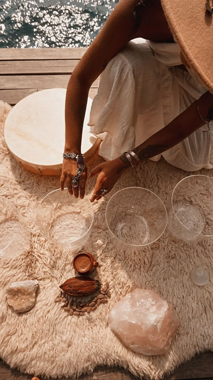 Pessoa usando vestido branco e chapéu marrom toca tigelas de cristal transparentes sobre tapete peludo ao lado de cacau, pedras e uma xícara de café.
