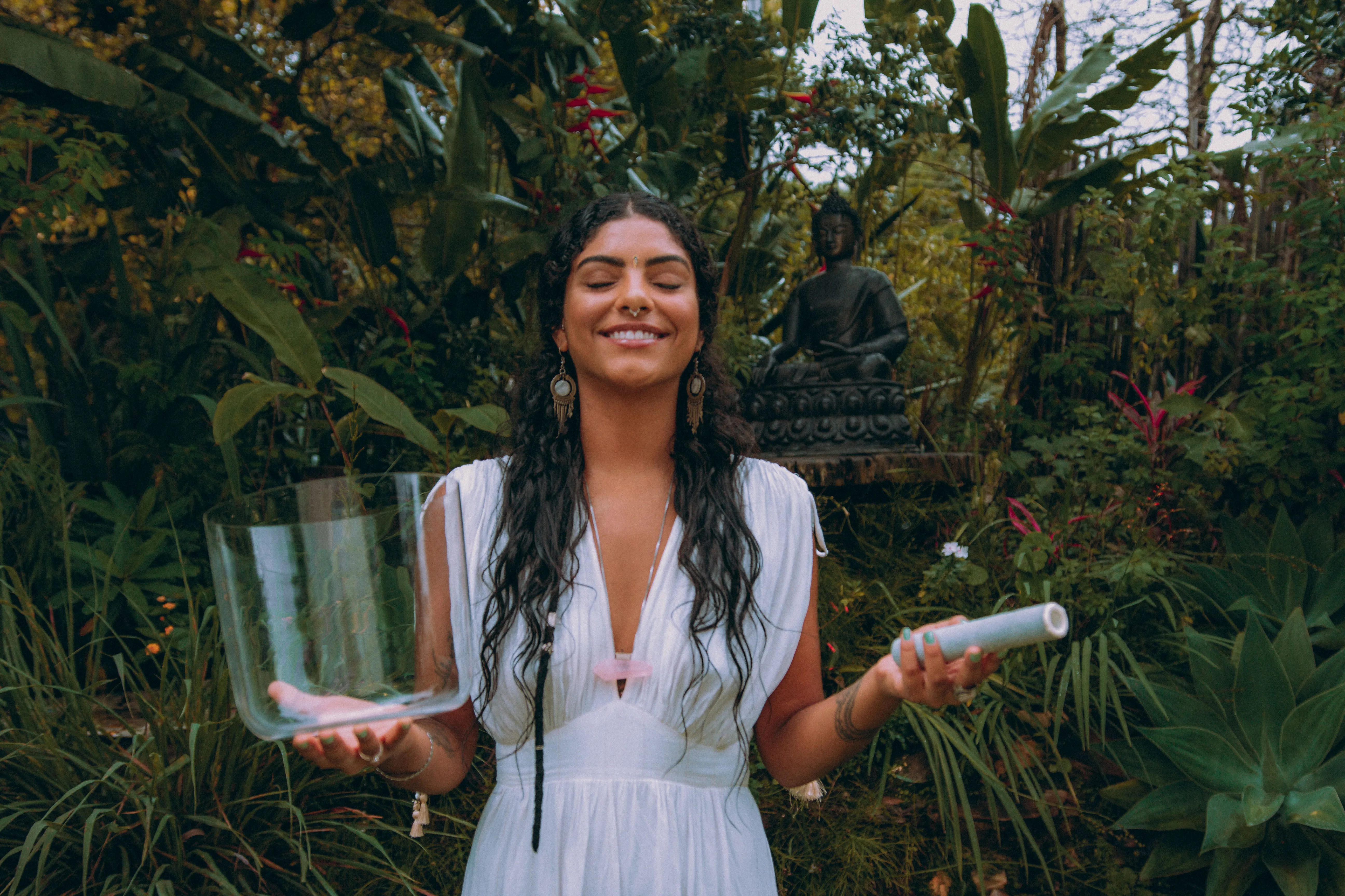 Mulher sorridente de vestido branco em um jardim com olhos fechados, segurando uma tigela de cristal em uma mão e um cilindro branco na outra.