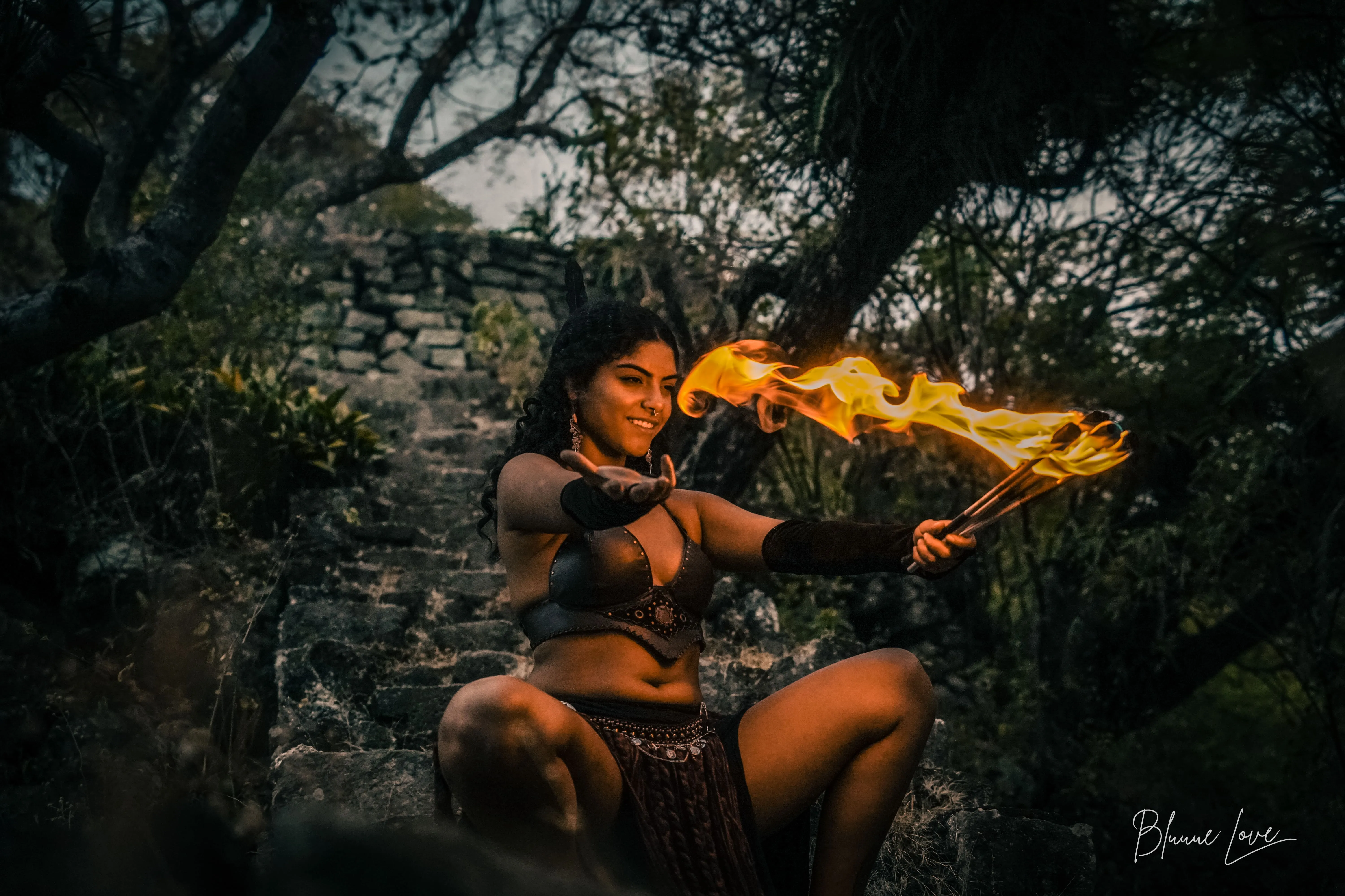 Mulher sentada em uma escadaria de pedra segurando tochas em chamas em um ambiente ao ar livre com árvores ao fundo.
