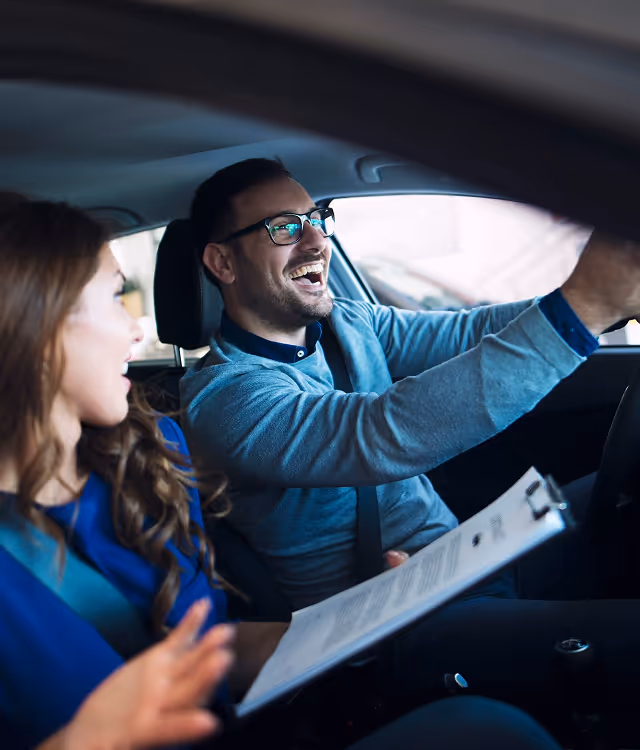 Man driving a car and smiling while a woman beside him holds a clipboard and looks on.