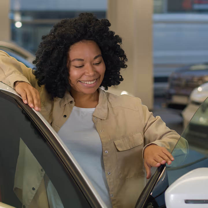 Smiling woman with curly hair opening the door of a white car in a showroom.