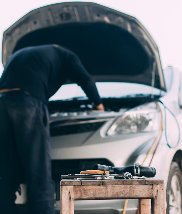 Car mechanic working under the open hood of a white car with tools on a wooden stool in the foreground.