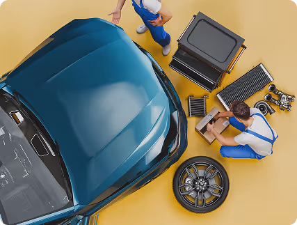 Two mechanics in blue overalls working near a blue car with car parts and tools spread on the floor.