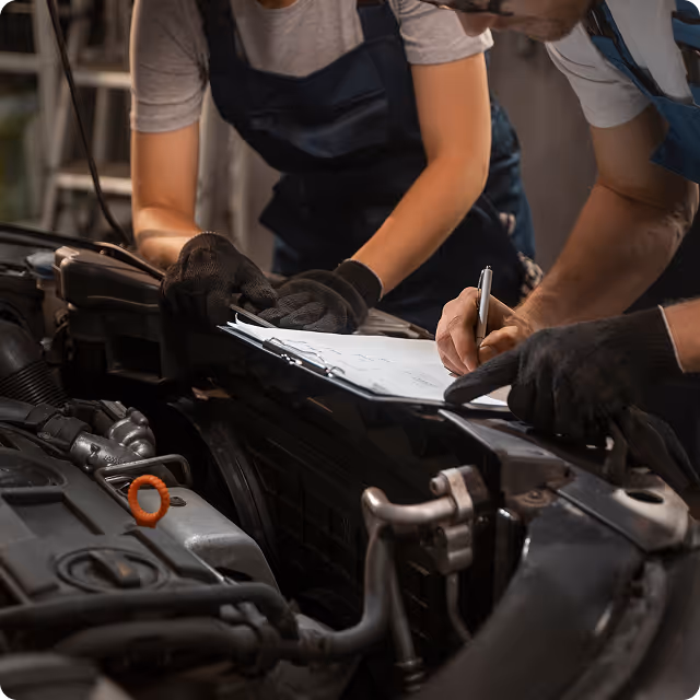 Two mechanics in gloves inspecting a car engine and documenting notes on a clipboard.