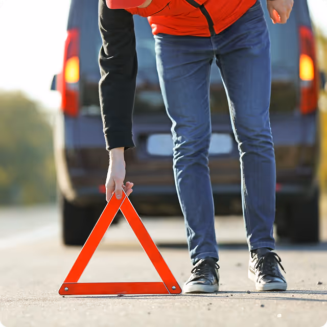 Person in red jacket placing a red emergency triangle on the road behind a dark vehicle.