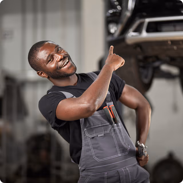 Smiling mechanic in overalls pointing upward in an auto repair shop.