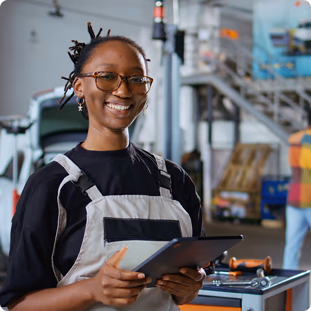 Smiling woman in work overalls holding a tablet in an industrial workshop.