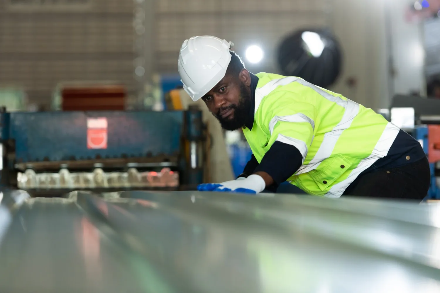 A metal worker in a hard hat and safety vest checks fabricated sheet metal in a factory.