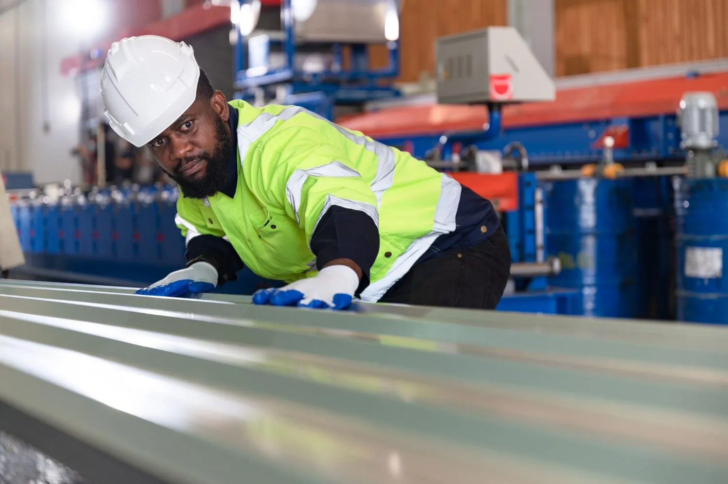 A man in a hard hat and high-visibility vest inspects corrugated sheet metal in a fabrication plant.