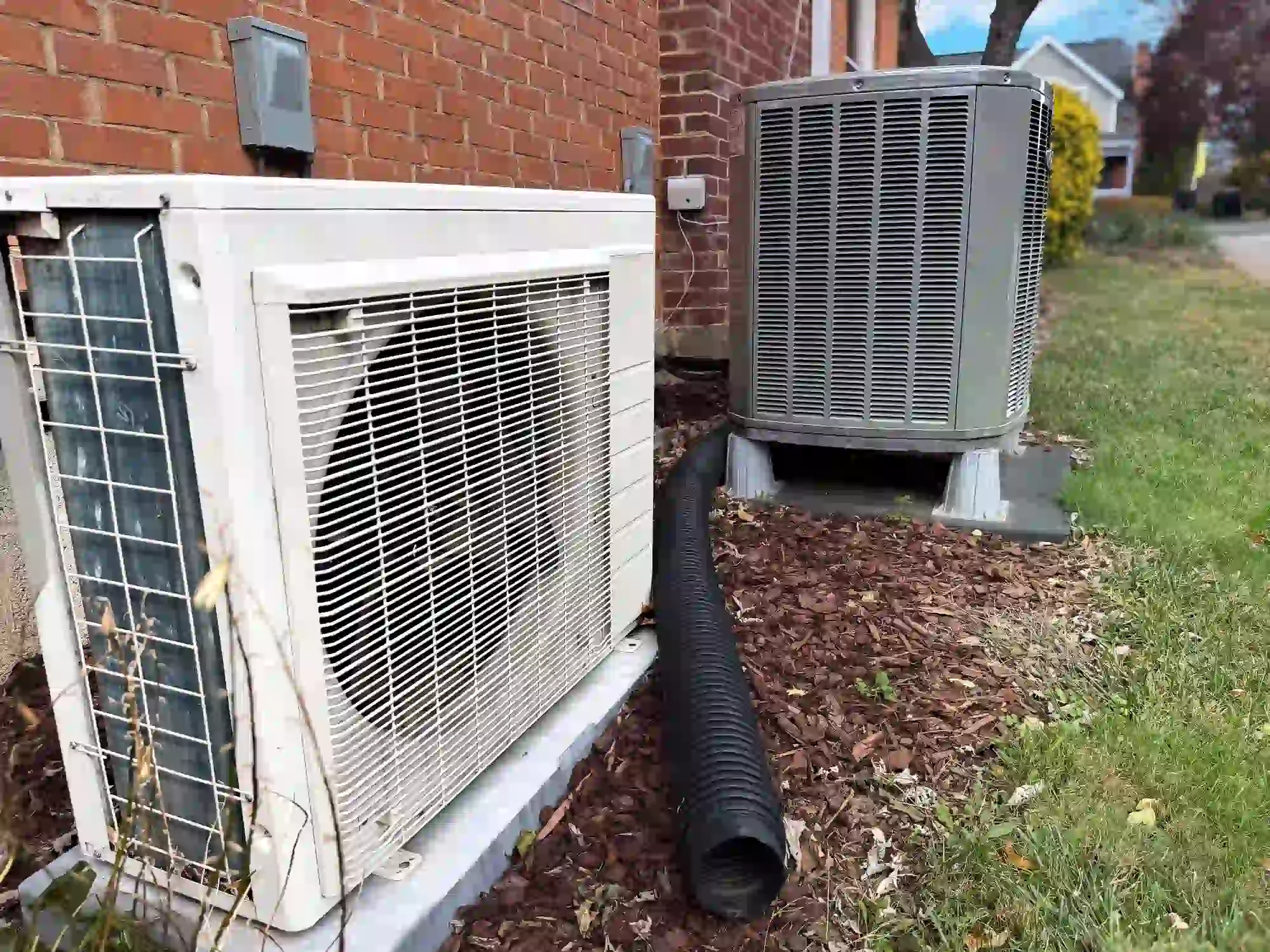  Two different HVAC condenser units sit side-by-side outside a red brick wall. The unit in the foreground is a smaller, white mini-split condenser, and the unit behind it is a larger, standard grey rectangular unit. A black corrugated drain pipe runs between them, over dark mulch and green grass.