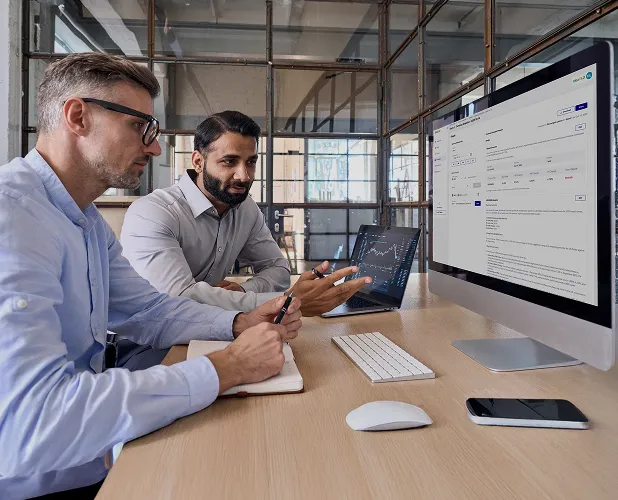 Two men in a modern office discussing data on a desktop computer and a laptop showing charts and text.