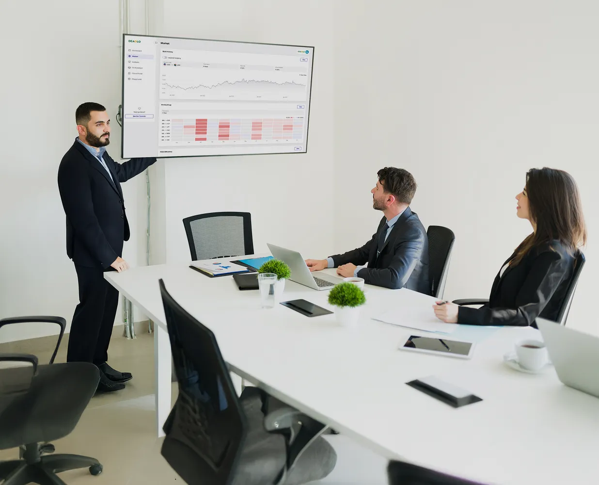 Man in suit presenting data charts on a wall-mounted screen to two colleagues seated at a conference table.