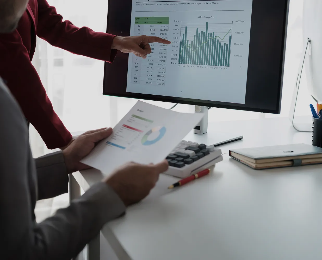 Two professionals analyzing financial charts and reports displayed on a computer screen and paper document.
