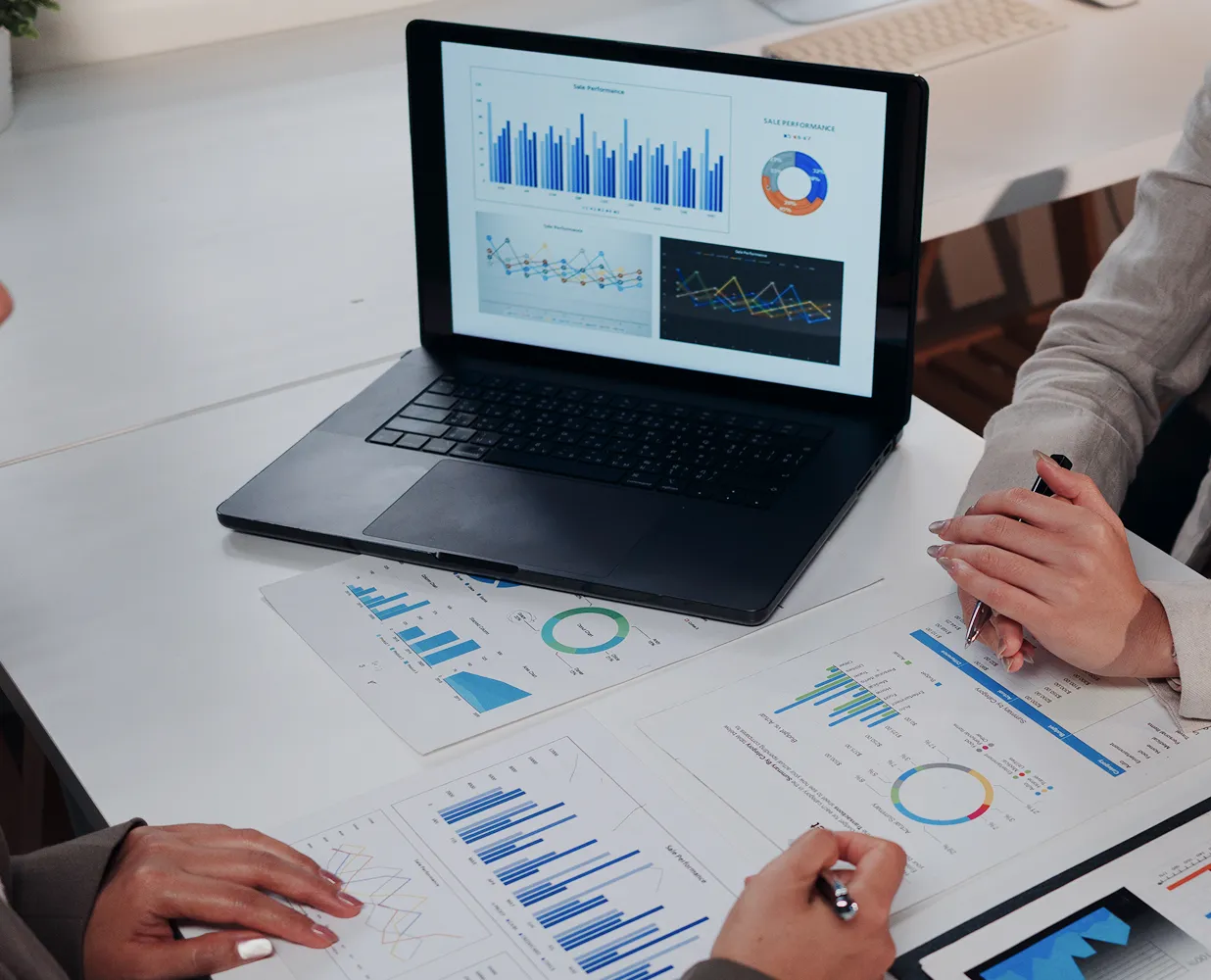 Two people reviewing printed sales and performance charts around a laptop displaying similar data graphs.