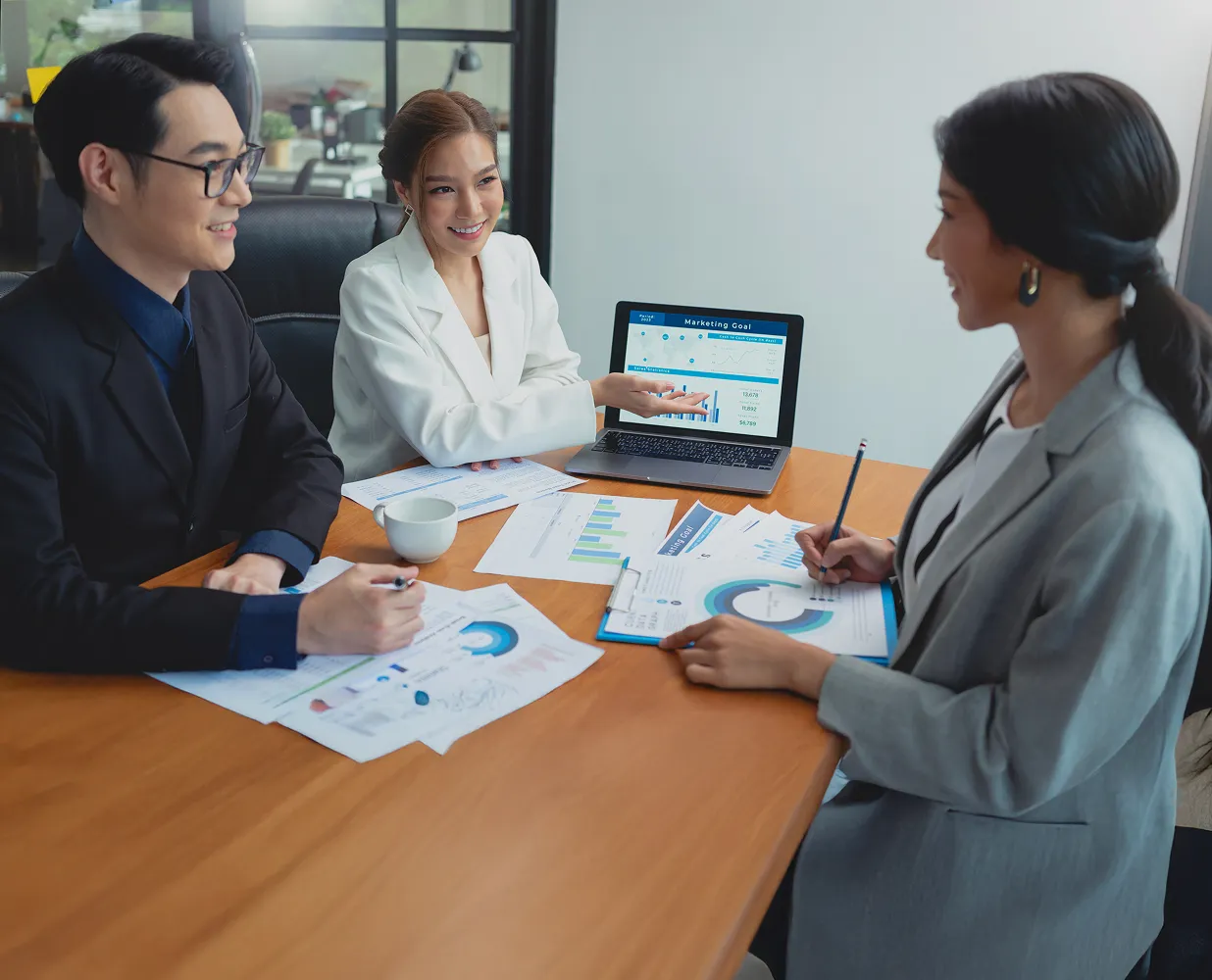 Three business professionals sitting around a table reviewing marketing goal charts on paper and a laptop during a meeting.