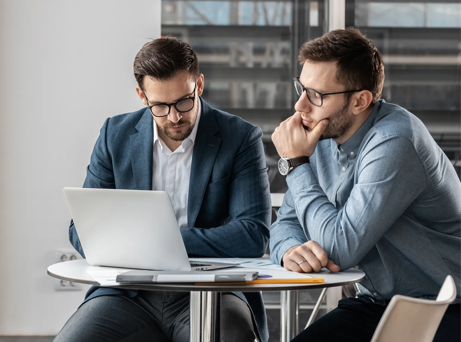Two men in glasses sitting at a round table, one working on a laptop while the other watches thoughtfully.