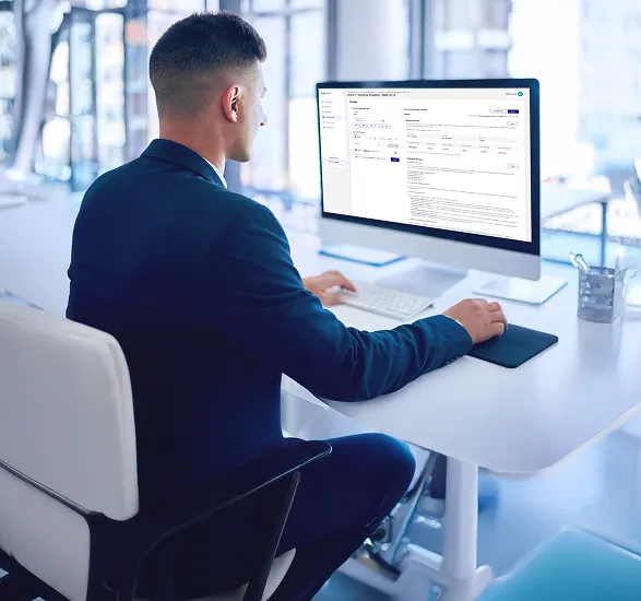 Man in a suit working on a desktop computer at a modern office desk with a web interface visible on the screen.
