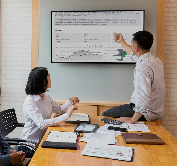 Two business colleagues in a meeting room reviewing financial graphs displayed on a wall screen.