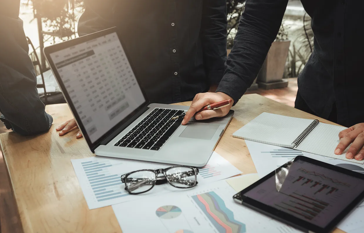 Person pointing at laptop screen with pen during a business meeting around a table with charts, glasses, and a tablet.