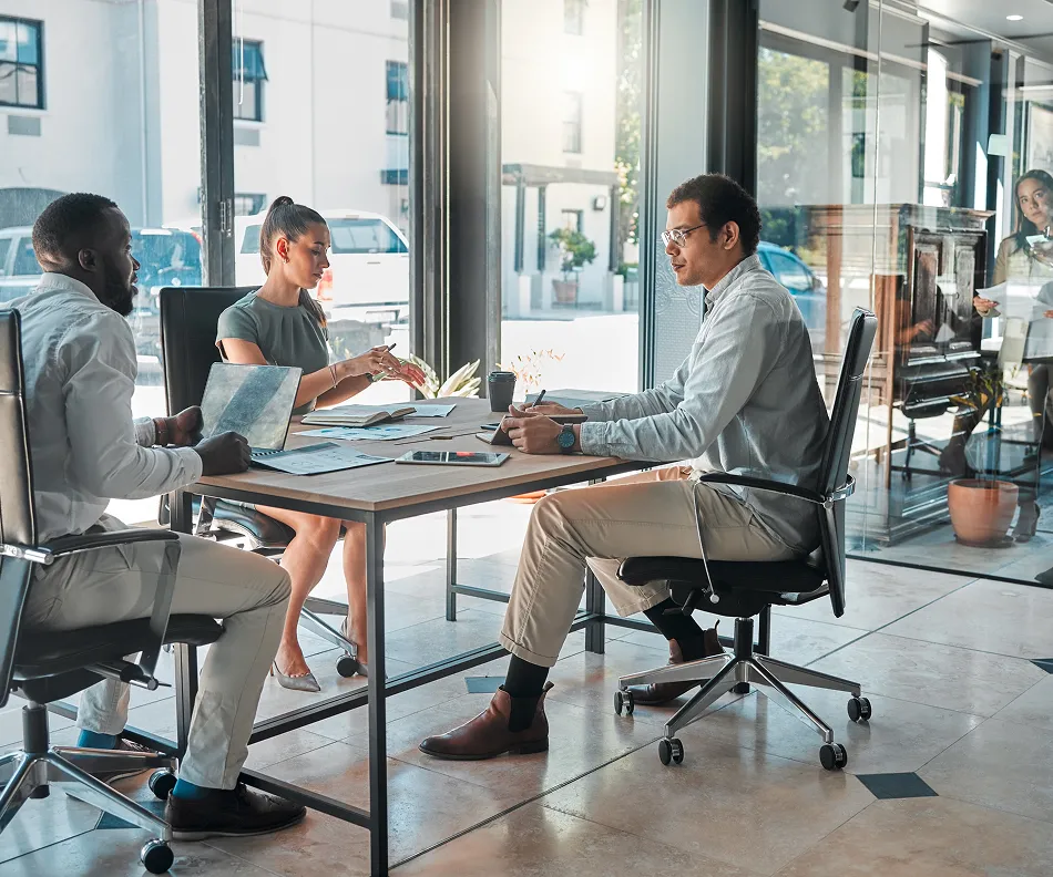 Three colleagues sitting around a table in a modern office, discussing work with laptops and documents, while a woman stands in the background holding papers.