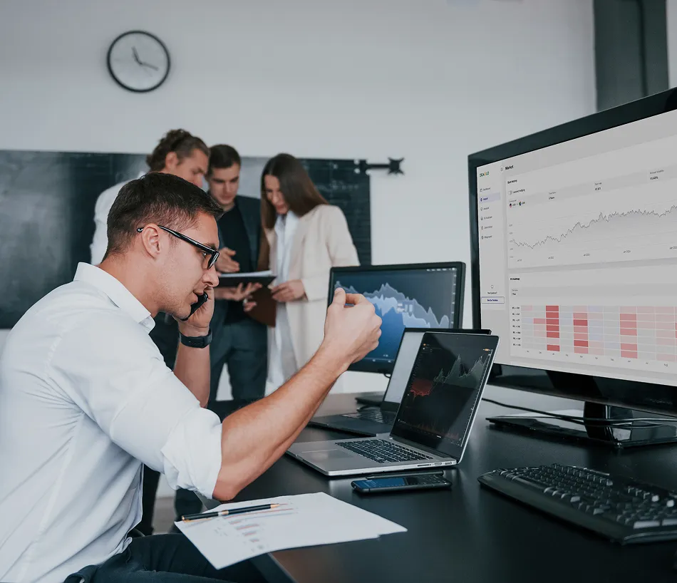 Man in glasses talking on phone at desk with multiple computer screens showing charts, while three colleagues discuss in the background.