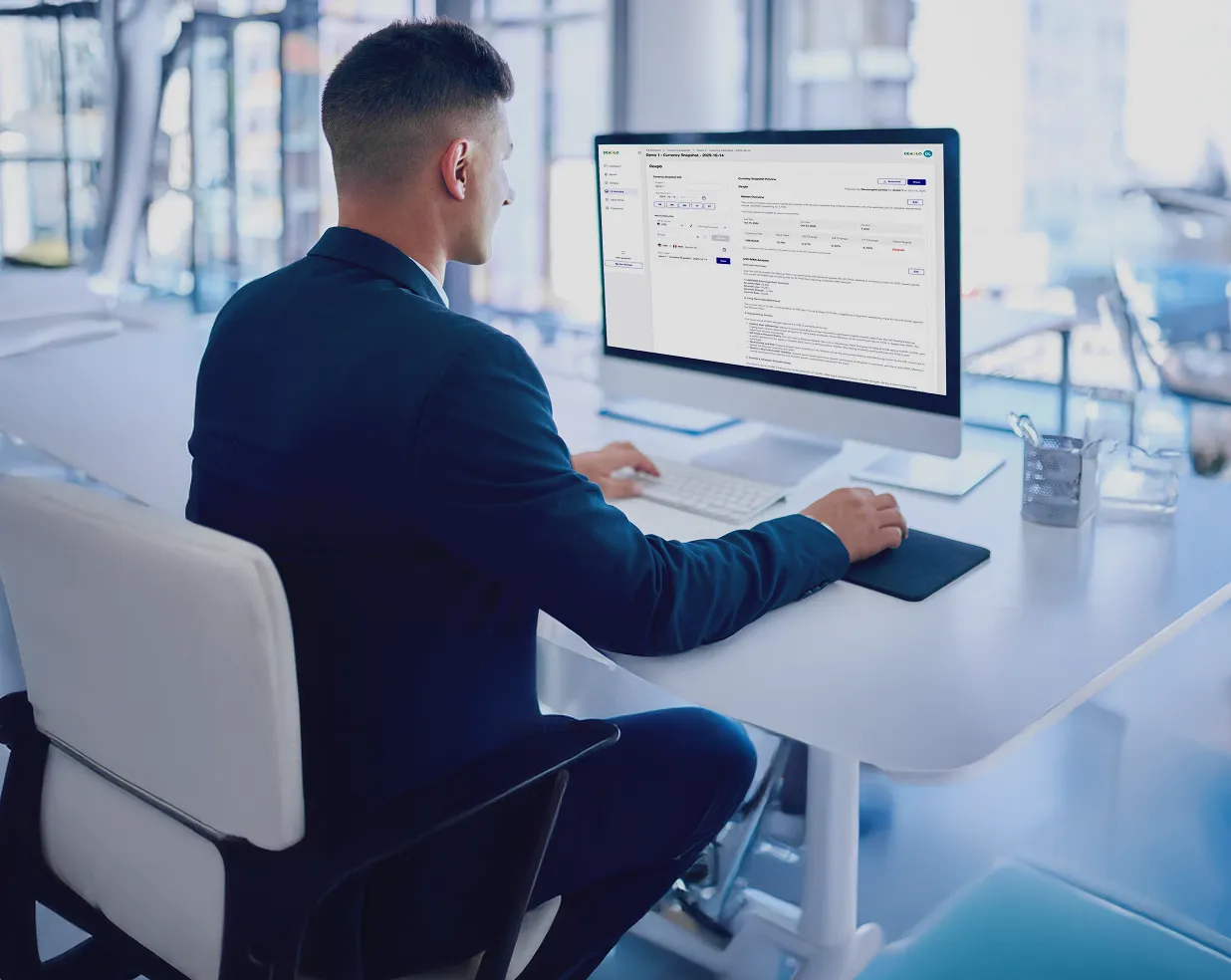 Man in a navy suit working on a desktop computer at a white office desk with a modern, bright office background.