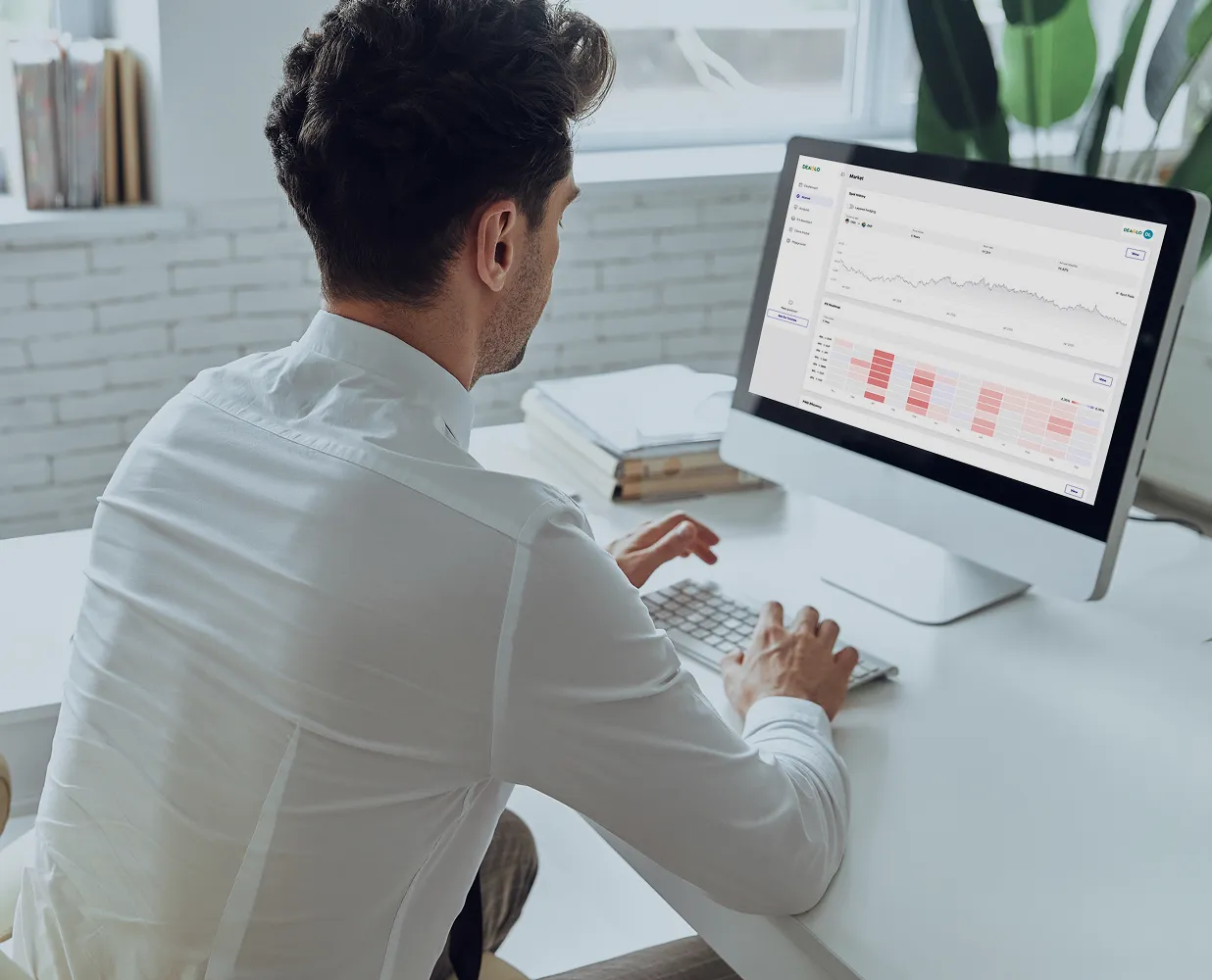 Man in white shirt typing on a keyboard in front of a desktop computer displaying market data charts.