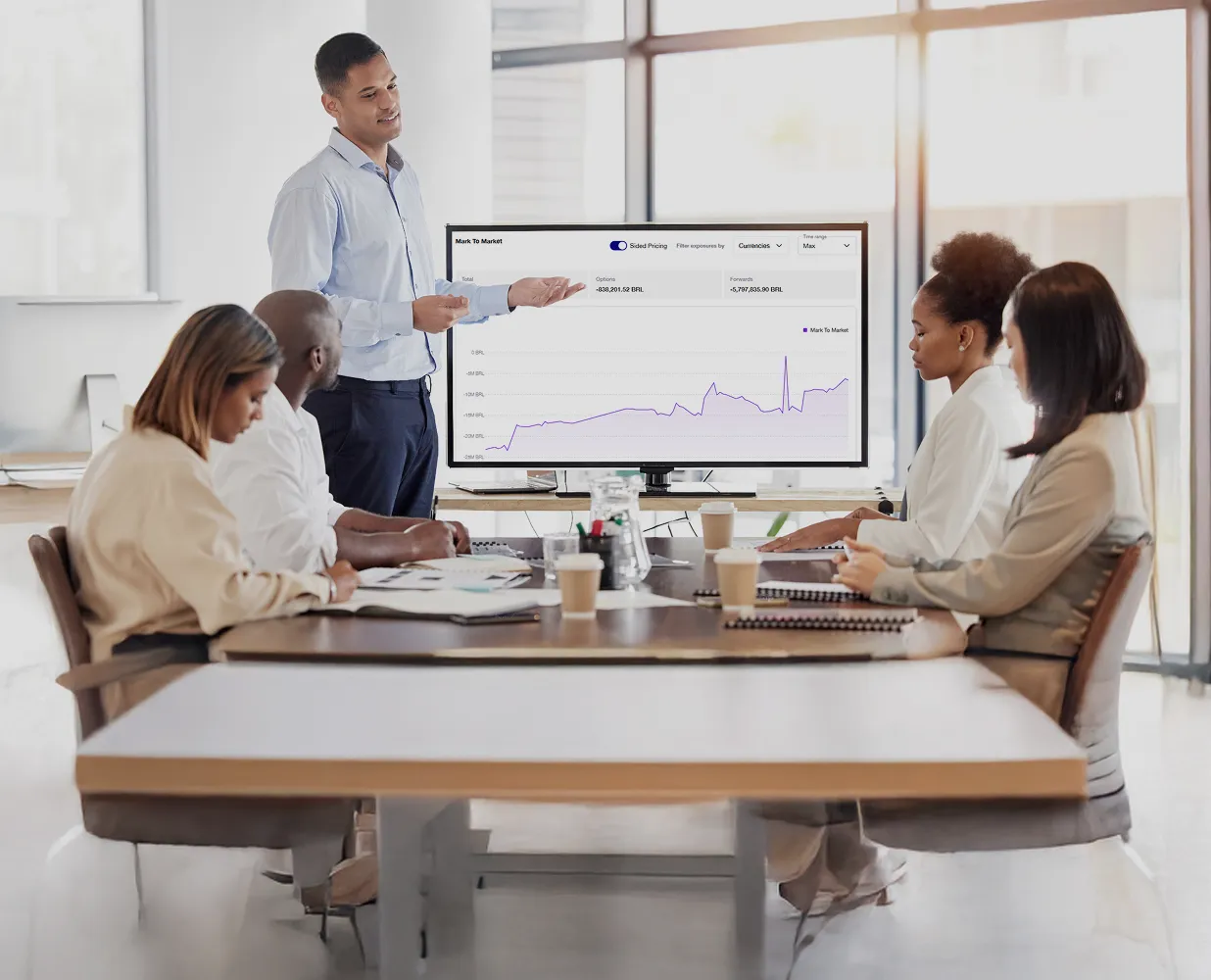 Businessman presenting financial chart on a monitor to four colleagues seated at a conference table in a bright office.