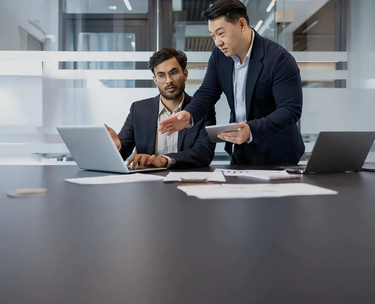 Two businessmen discussing work at a table with laptops and papers in a modern office.