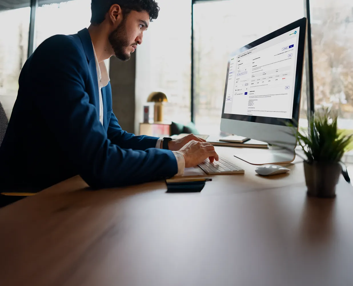Man in blue blazer working on an iMac at a desk in a modern office with large windows.