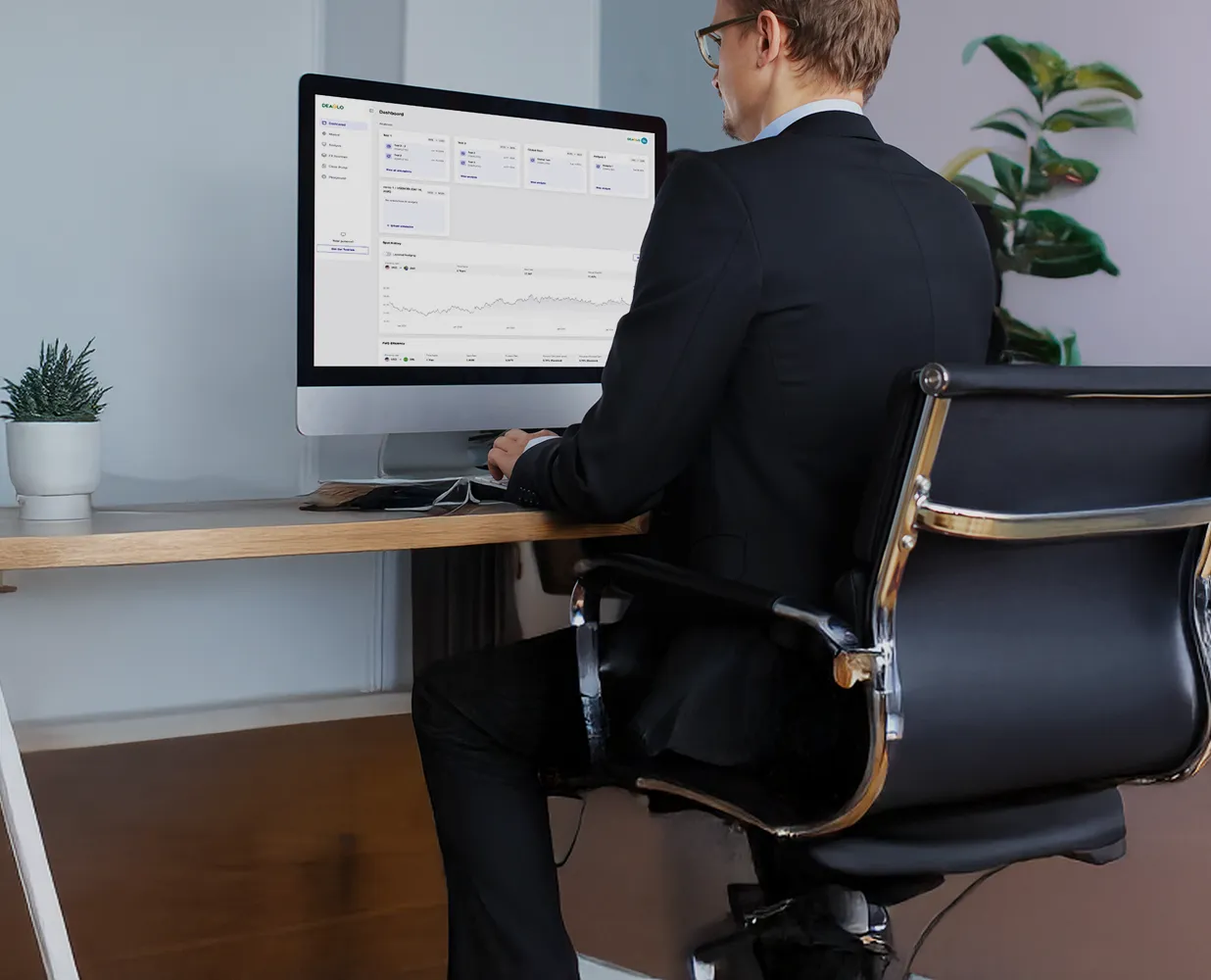 Man in a black suit sitting at a desk working on a computer with a dashboard displaying charts and data.