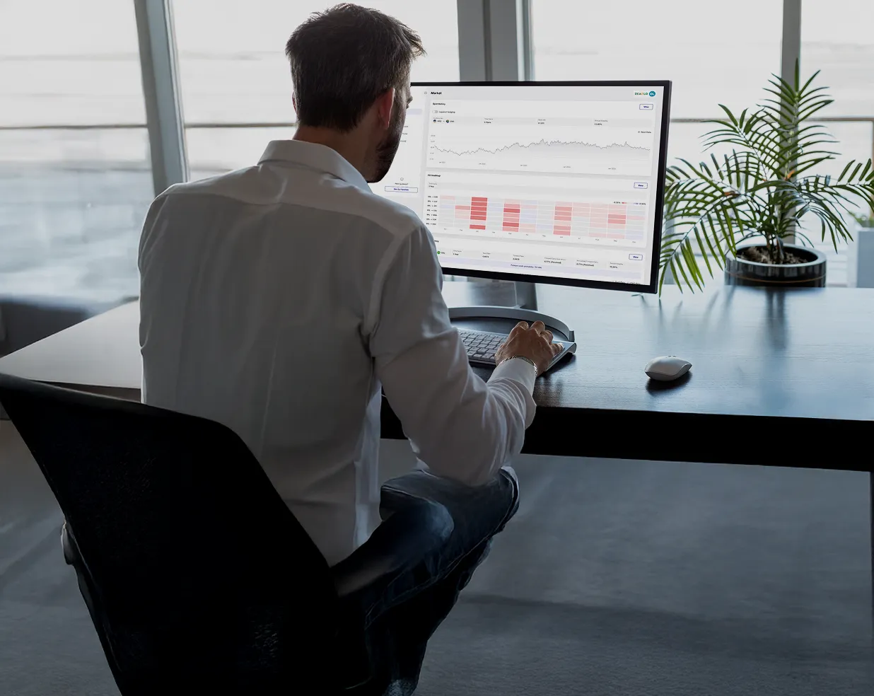 Man in white shirt sitting at desk working on computer with financial charts and graphs on screen.