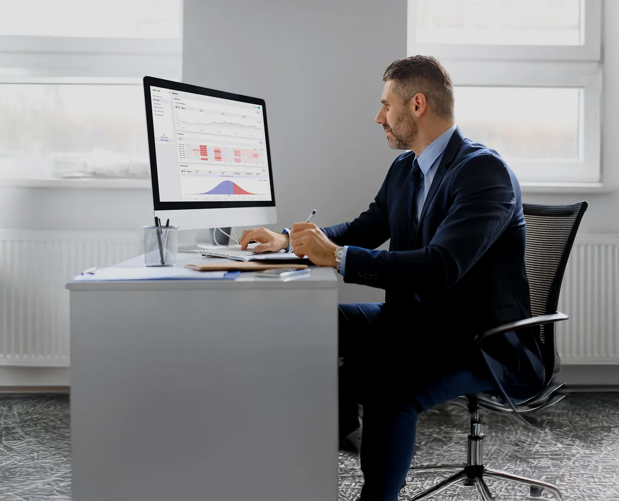 Man in a suit sitting at a desk analyzing financial data on a desktop computer in a bright office.