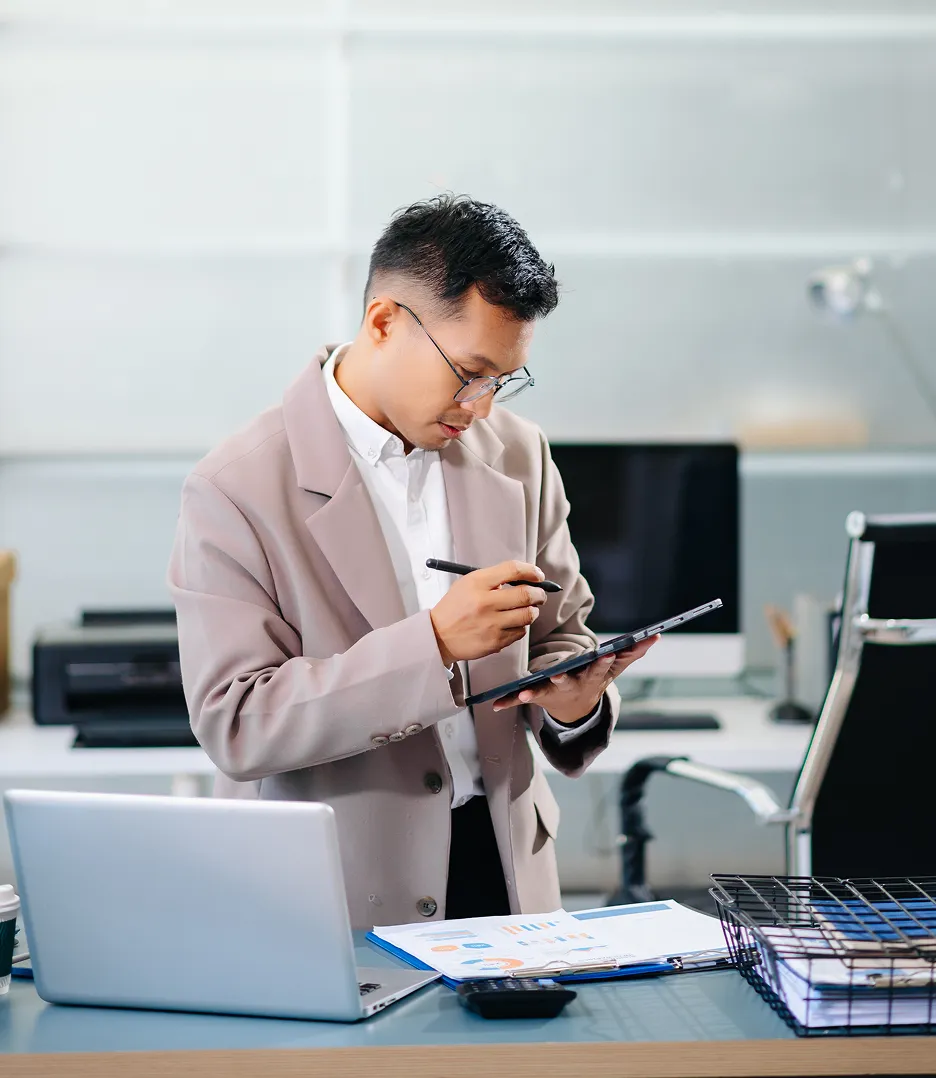 Man wearing glasses and a beige blazer using a digital pen on a tablet in an office setting with a laptop and documents on the desk.