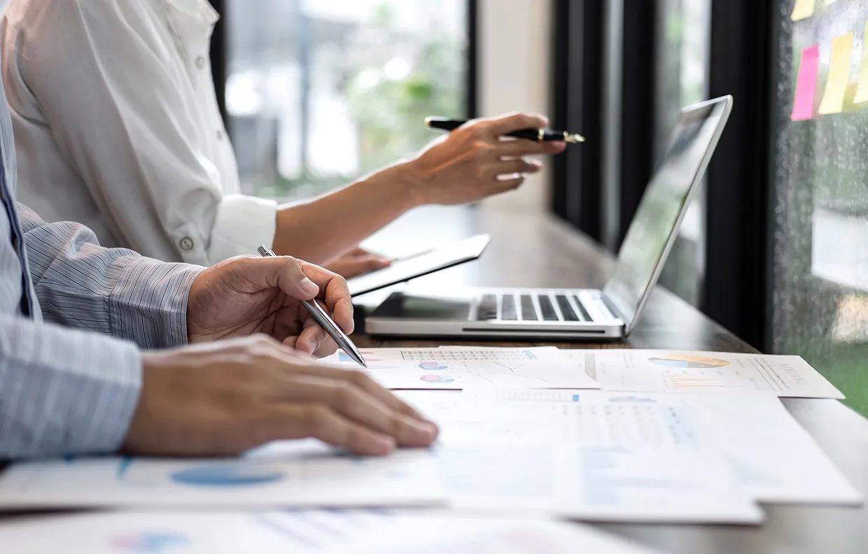 Two people working at a desk with charts and a laptop, one holding a pen and pointing at documents.