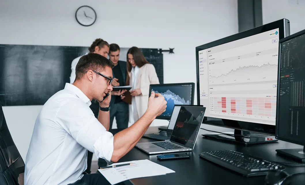 Man in white shirt talking on phone and gesturing in front of multiple computer screens displaying charts, with three colleagues discussing in background.