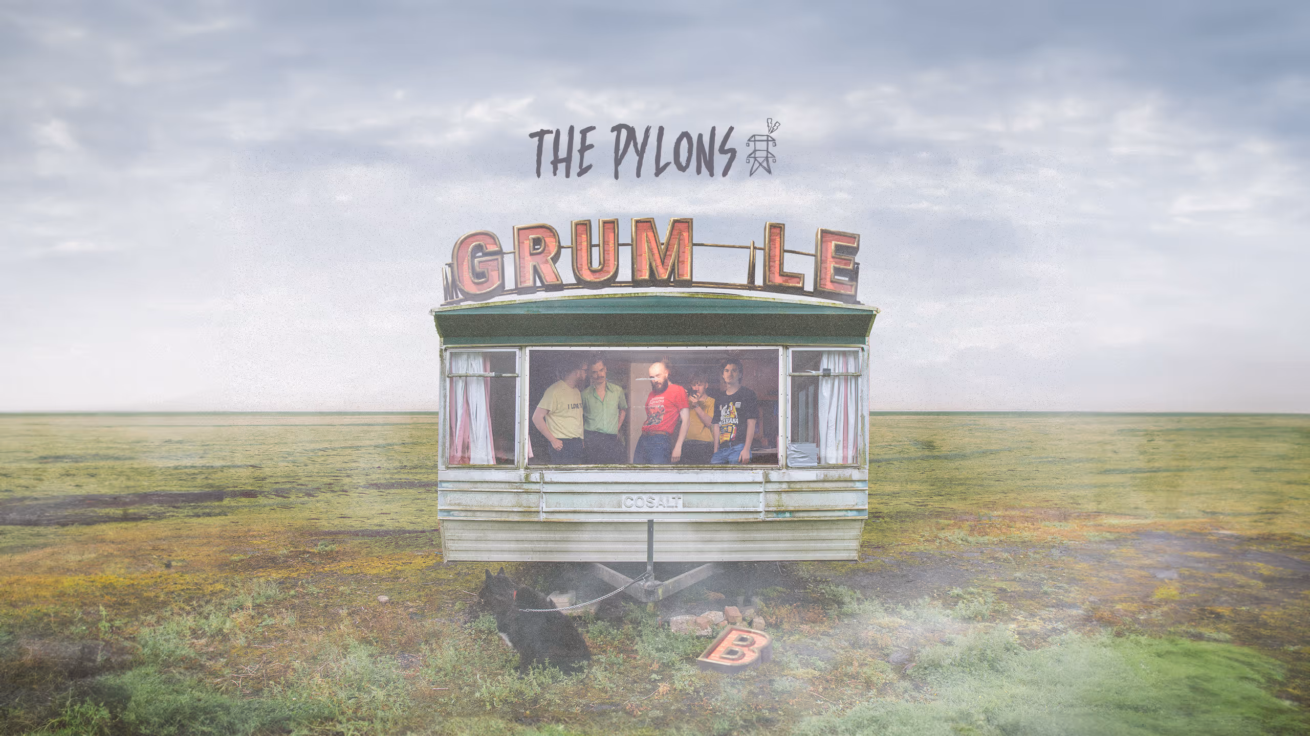 Five men standing inside a small green trailer with the letters 'GRUM LE' on top and a black dog tied to the trailer in a wide grassy field.