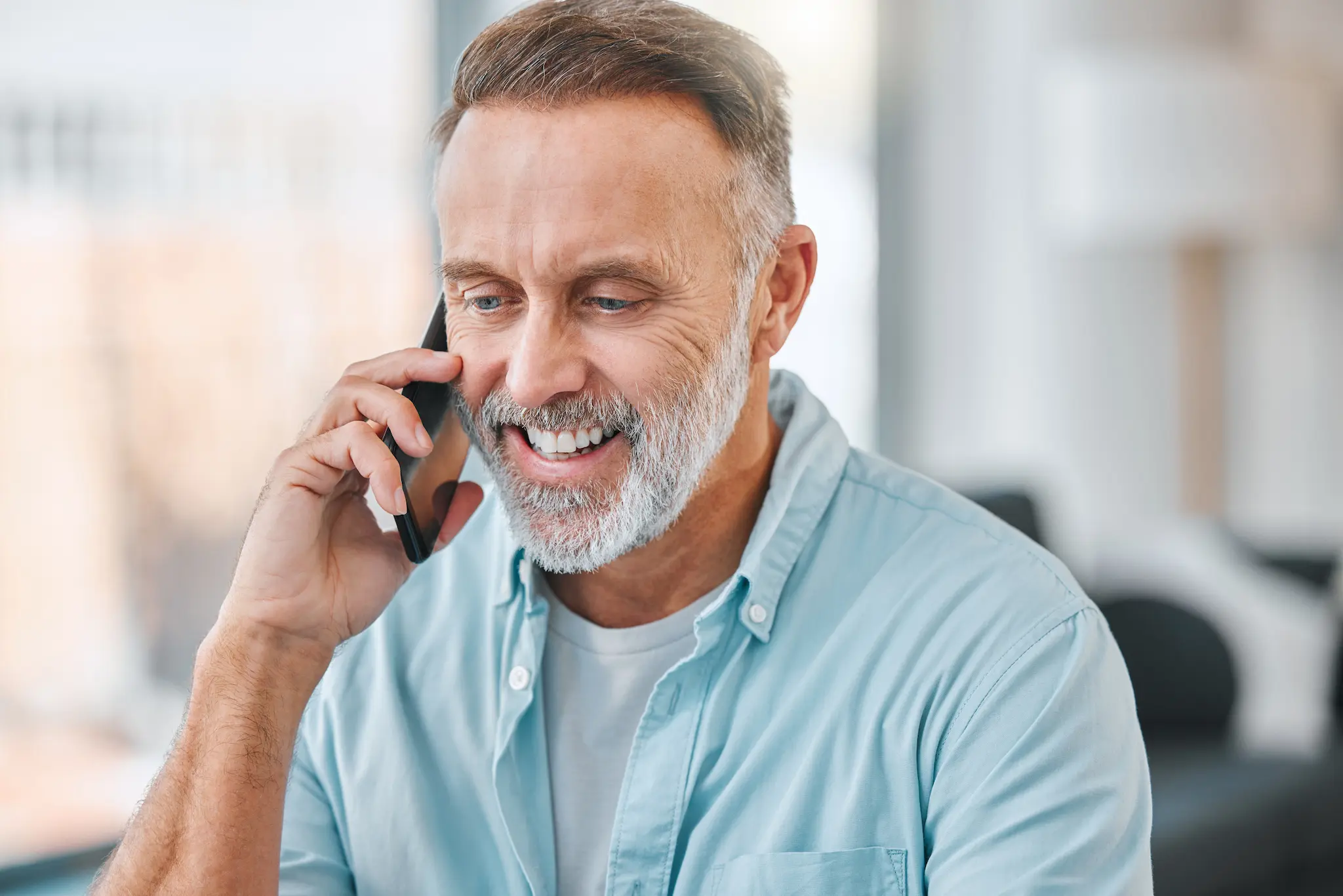 Smiling middle-aged man with gray beard talking on a smartphone in a bright room.