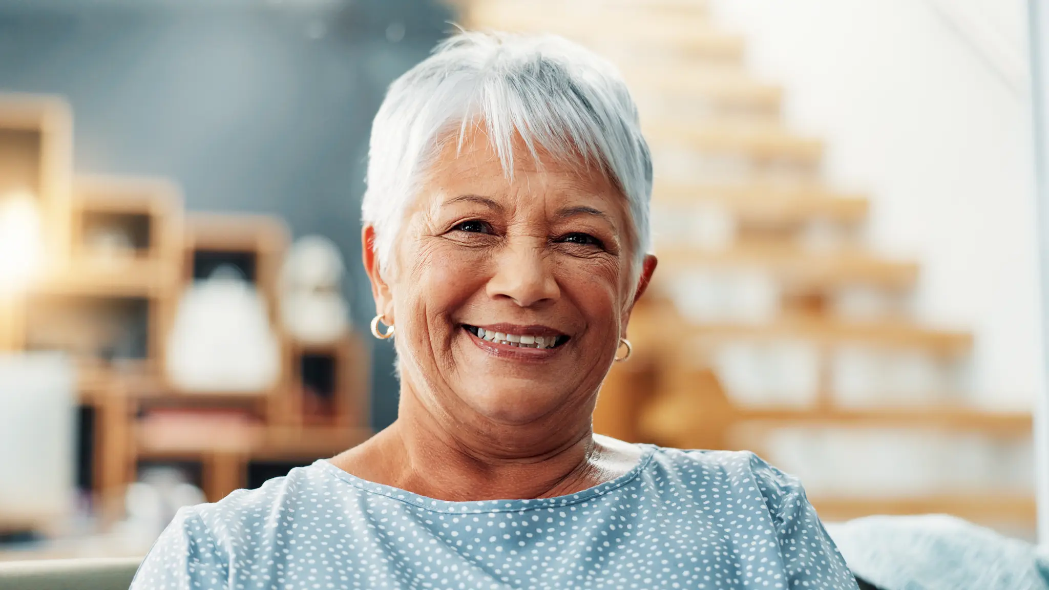 Smiling older woman with short white hair wearing a blue polka dot blouse, sitting indoors with blurred background.