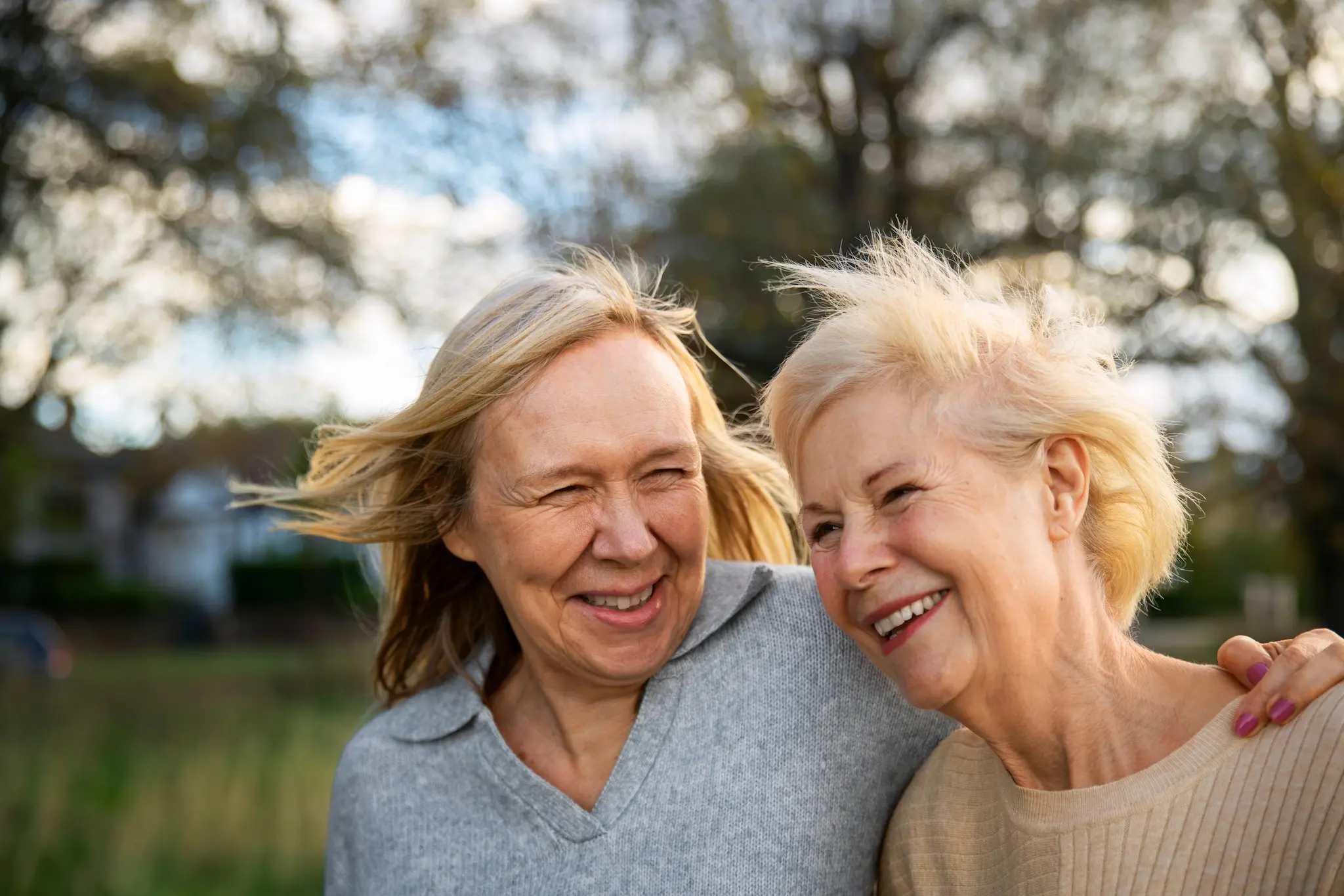 Two smiling older women outdoors with wind blowing their hair, sharing a joyful moment.