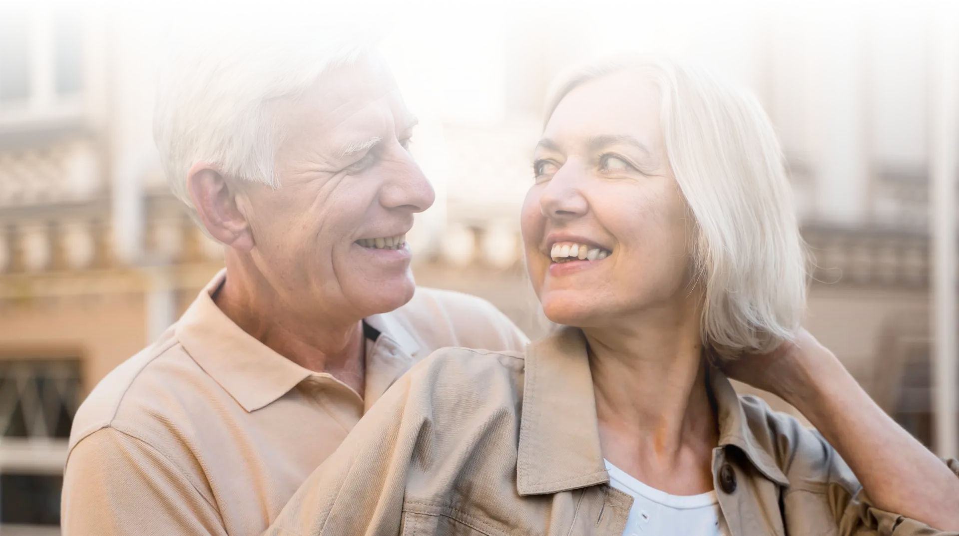 Smiling elderly couple looking at each other outdoors with blurred building background.