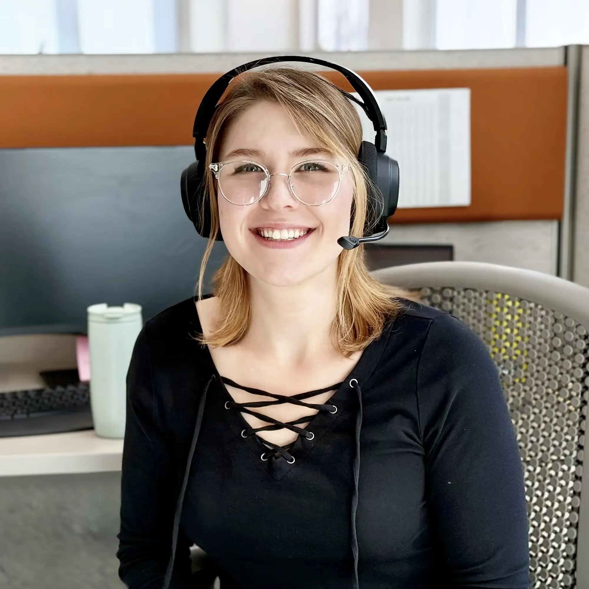 Smiling woman wearing glasses and a headset sitting at an office desk with a computer and a tumbler.