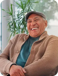 Elderly man wearing a cap and brown sweater smiling while sitting indoors.