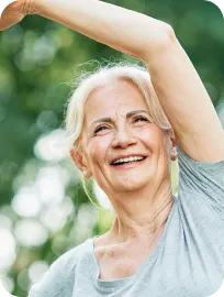 Smiling elderly woman raising her arm outdoors with greenery in the background.
