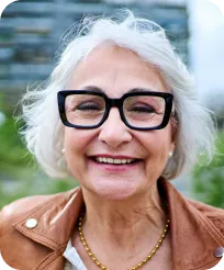 Smiling elderly woman with white hair, black glasses, and a brown jacket outdoors.